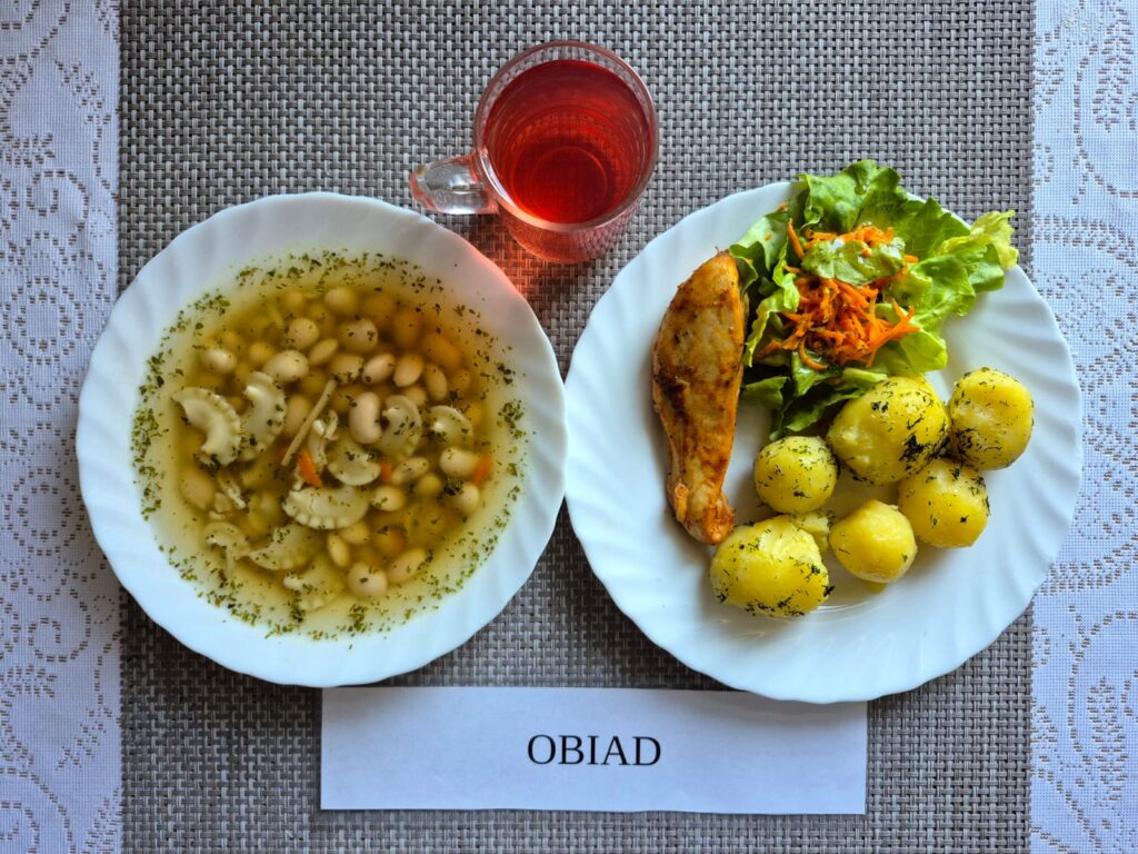 Na zdjęciu znajduję się posiłek w którego skład wchodzi Zupa fasolowa z makaronem, Filet z kurczaka pieczony, Ziemniaki, Sałata masłowa z marchewką oraz Kompot z jabłek