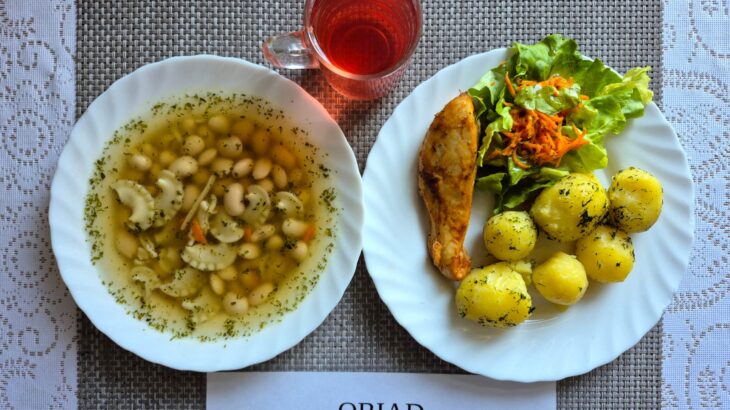 Na zdjęciu znajduję się posiłek w którego skład wchodzi Zupa fasolowa z makaronem, Filet z kurczaka pieczony, Ziemniaki, Sałata masłowa z marchewką oraz Kompot z jabłek