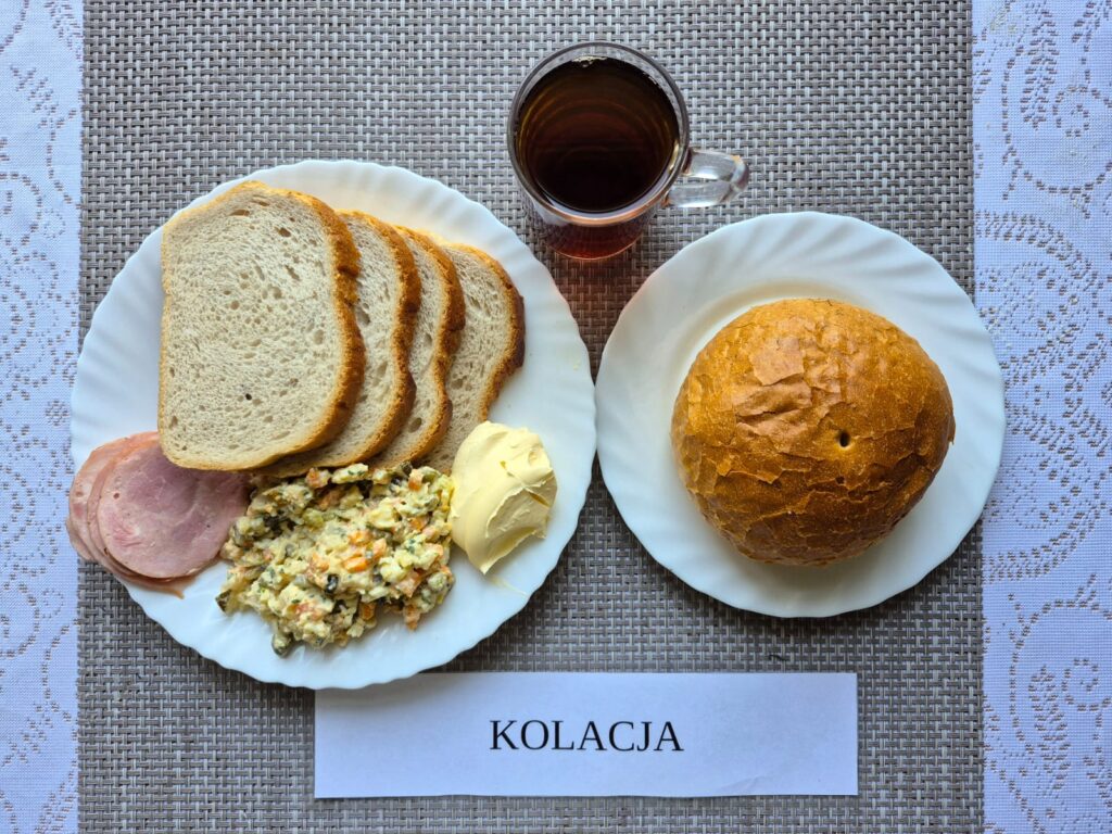 Na zdjęciu znajduję się posiłek w którego skład wchodzi Chleb pszenny, Margaryna Śniadaniowa, Sałatka jarzynowa z młodych jarzyn, bułka pszenna, wędlina oraz Herbata czarna