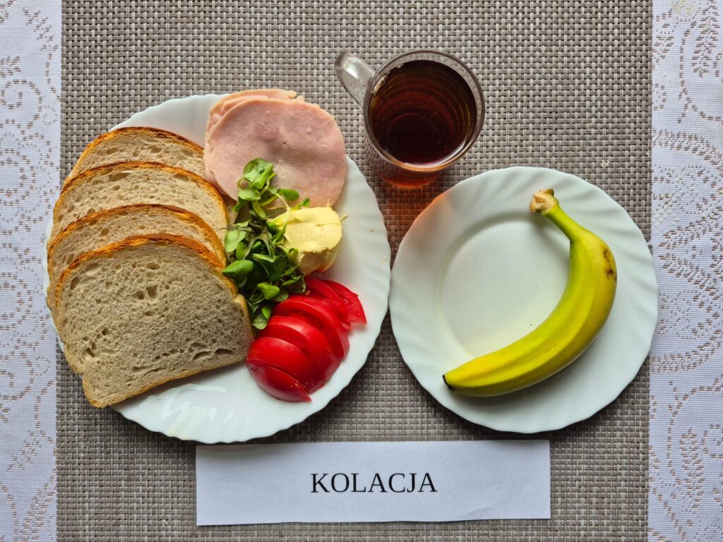 Na zdjęciu znajduję się posiłek w którego skład wchodzi Chleb pszenny, Margaryna Śniadaniowa, Szynka konserwowa, Rukola, Pomidor, banan oraz Herbata czarna