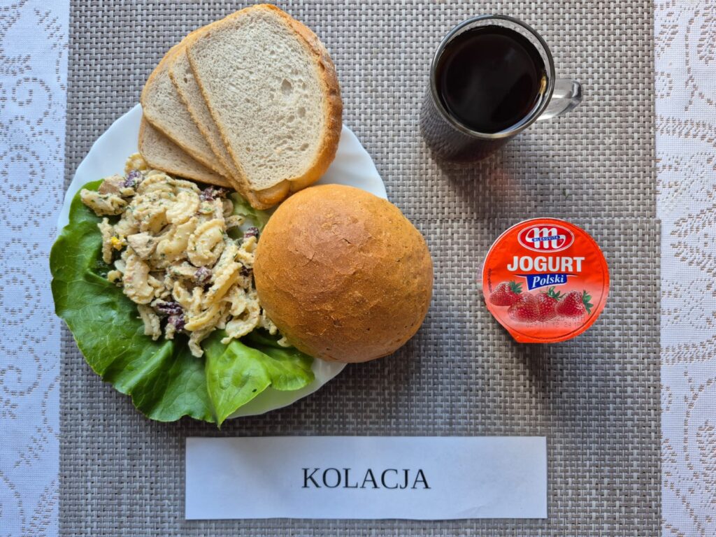 Na zdjęciu znajduję się posiłek w którego skład wchodzi Chleb pszenny, Bułka pszenna, Margaryna śniadaniowa, Sałatka z kurczakiem i fetą z dodatkiem kaszy kuskus, jogurt oraz Herbata czarna