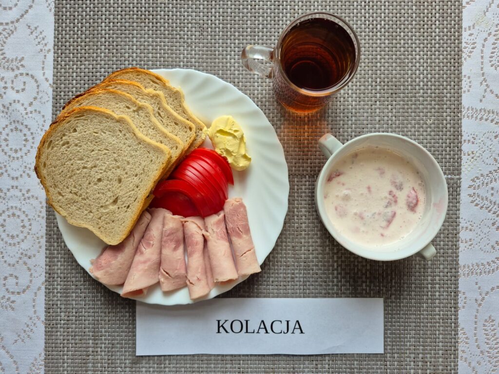 Na zdjęciu znajduję się posiłek w którego skład wchodzi Chleb pszenny, Margaryna Śniadaniowa, Kiełbasa krakowska, Pomidor oraz Herbata czarna