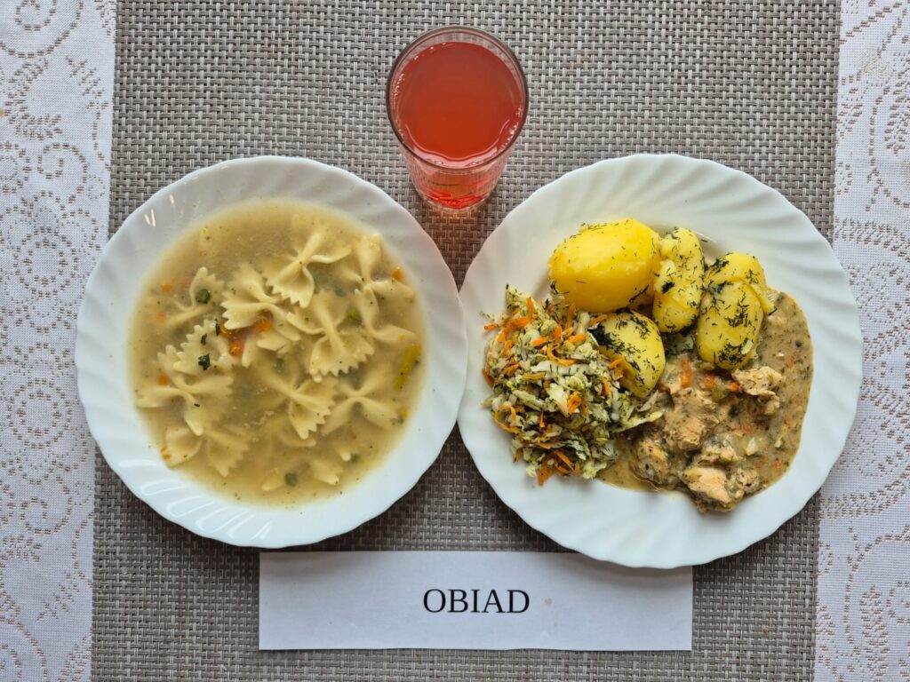 Na zdjęciu znajduję się posiłek w którego skład wchodzi Zupa jarzynowa z makaronem, Ziemniaki, Filet z kurczaka zapiekany w sosie porowym, Surówka z kapusty pekińskiej i marchewki oraz kompot
