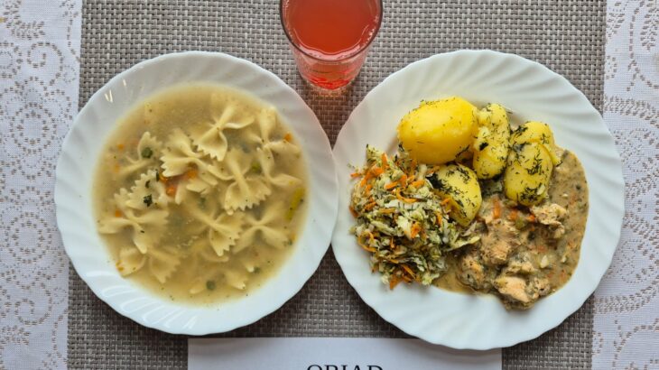 Na zdjęciu znajduję się posiłek w którego skład wchodzi Zupa jarzynowa z makaronem, Ziemniaki, Filet z kurczaka zapiekany w sosie porowym, Surówka z kapusty pekińskiej i marchewki oraz kompot