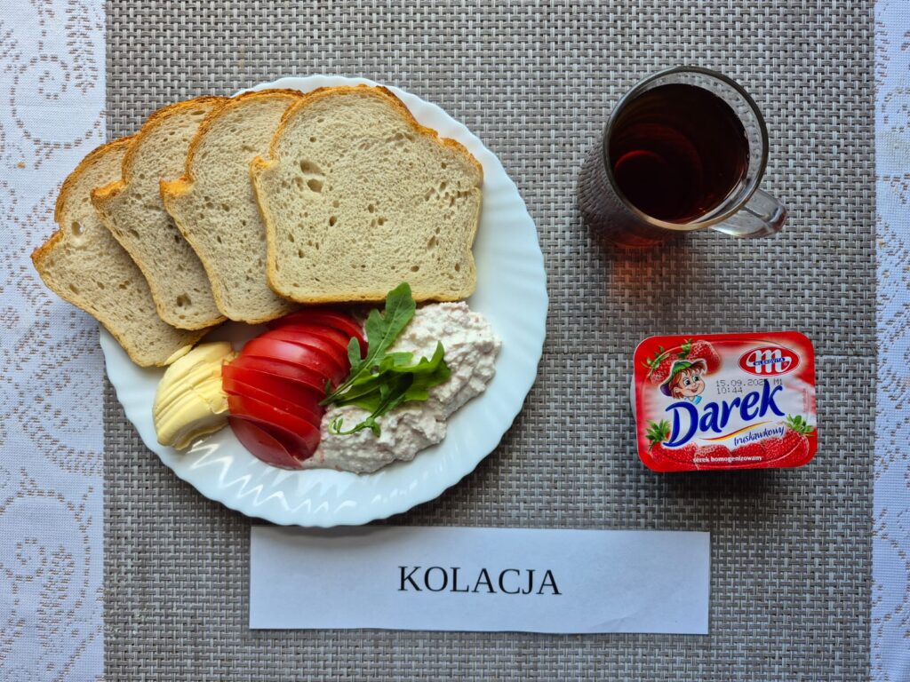 Na zdjęciu znajduję się posiłek w którego skład wchodzi Chleb pszenny, margaryna śniadaniowa, Pasta z twarogu i wędliny, Rukola, pomidor, serek oraz Herbata czarna