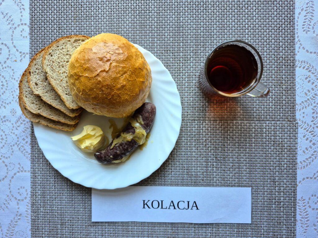 Na zdjęciu znajduję się posiłek w którego skład wchodzi Chleb pszenny, Margaryna Śniadaniowa, Kiszka pieczona z cebulą, bułka oraz Herbata czarna