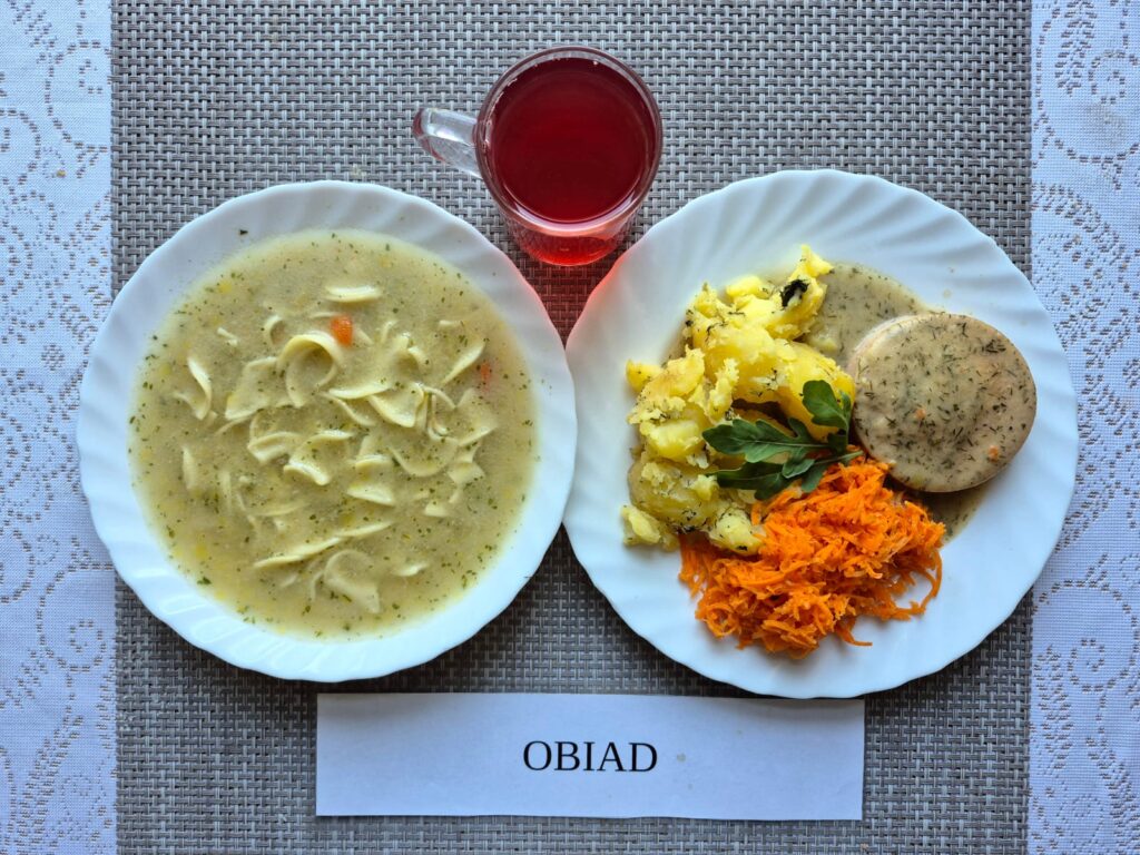 Na zdjęciu znajduję się posiłek w którego skład wchodzi Zupa brokułowa z makaronem, Ziemniaki, Bitka wieprzowa w sosie własnym, Sałatka z pomidora z cebulą oraz Kompot z jabłek