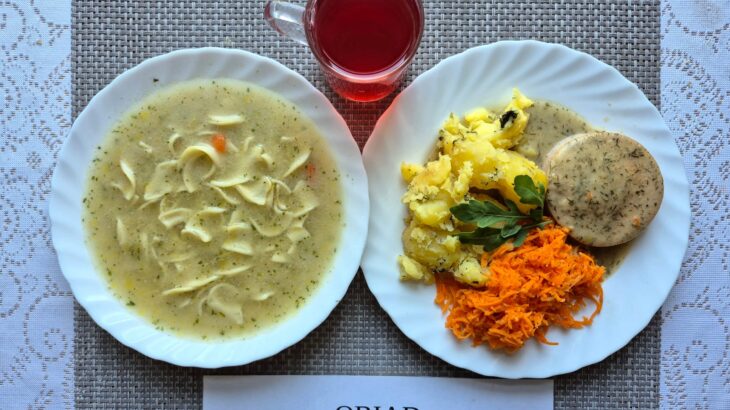 Na zdjęciu znajduję się posiłek w którego skład wchodzi Zupa brokułowa z makaronem, Ziemniaki, Bitka wieprzowa w sosie własnym, Sałatka z pomidora z cebulą oraz Kompot z jabłek