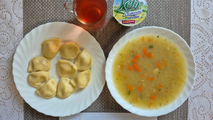 Na zdjęciu znajduję się posiłek w którego skład wchodzi Zupa ryżowa, Ziemniaki, Pierogi, kefir oraz kompot z jabłek
