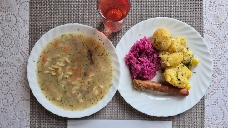 Na zdjęciu znajduję się posiłek w którego skład wchodzi Zupa pieczarkowa z makaronem, Ziemniaki, Kiełbasa pieczona, Surówka z czerwonej kapusty oraz Kompot z jabłek