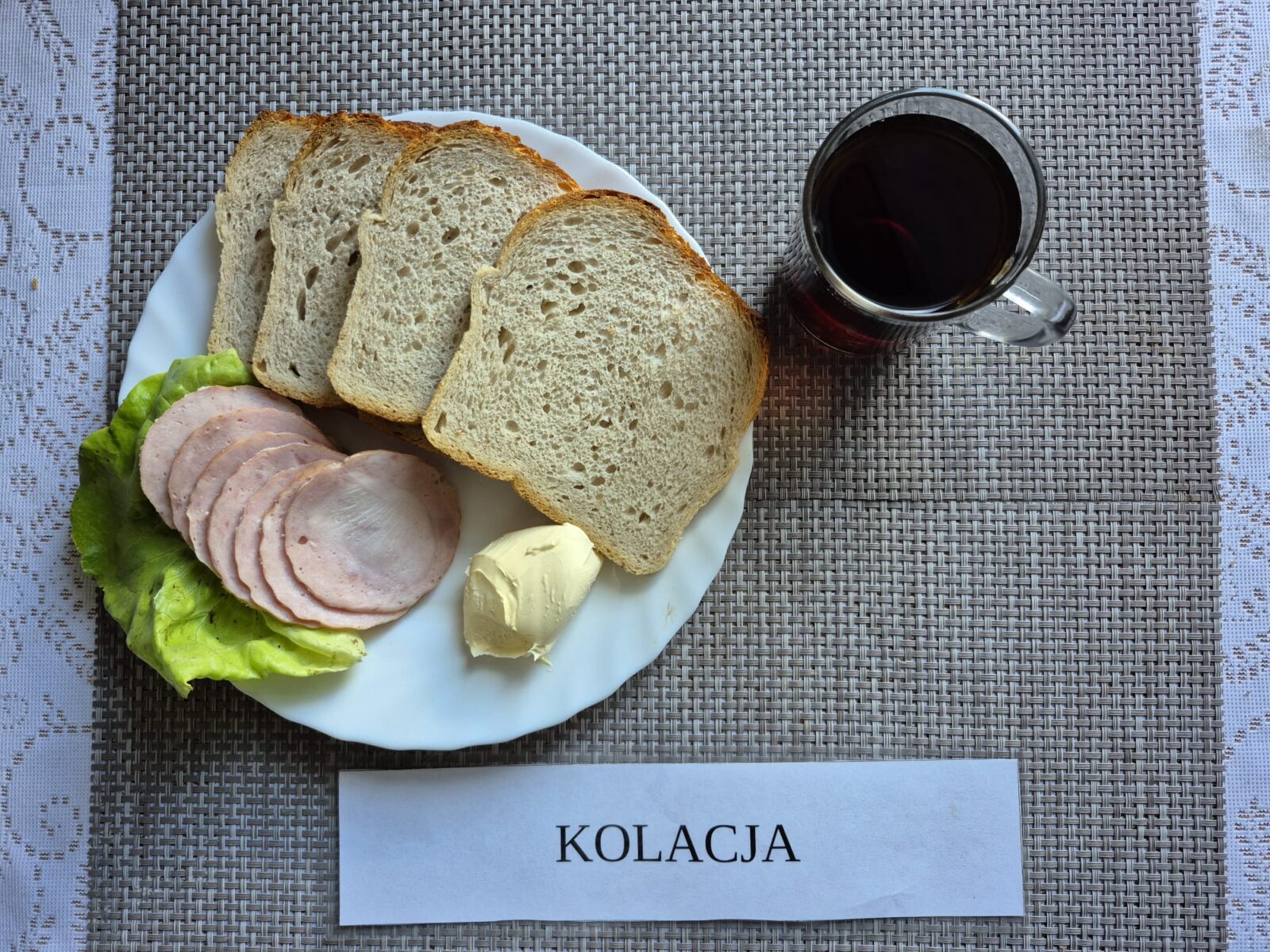 Na zdjęciu znajduję się posiłek w którego skład wchodzi Chleb pszenny, Margaryna Śniadaniowa, Kiełbasa krakowska drobiowa drobiowa, Sałata masłowa oraz Herbata czarna