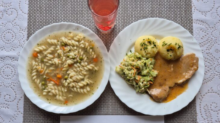 Na zdjęciu znajduję się posiłek w którego skład wchodzi Zupa jarzynowa z groszkiem cukrowym i makaronem 300 ml Ziemniaki 300g karkówka pieczona w sosie własnym 140g Surówka z ogórka zielonego i kapusty pekińskiej i marchewki oraz Kompot z jabłek