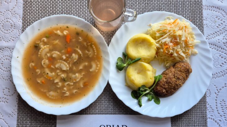 Na zdjęciu znajduję się posiłek w którego skład wchodzi Zupa ogonowa z makaronem, Sznycel smażony, Ziemniaki, Surówka z kapusty białej, z zalewą octową oraz Kompot z jabłek