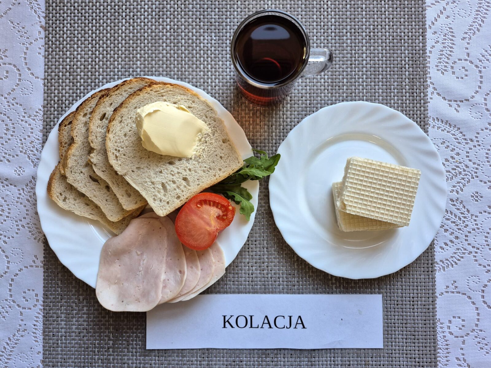 Na zdjęciu znajduję się posiłek w którego skład wchodzi Chleb pszenny, Margaryna Śniadaniowa, Kiełbasa krakowska drobiowa, pomidor oraz Herbata czarna