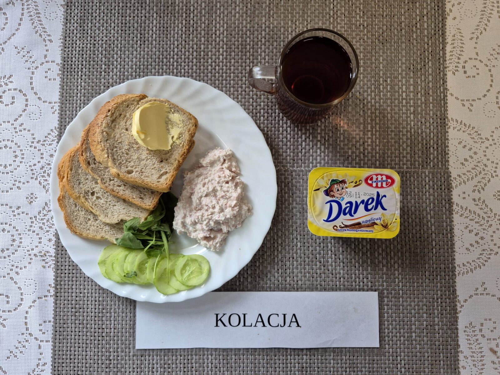 Na zdjęciu znajduję się posiłek w którego skład wchodzi Chleb razowy, Chleb pszenny, Margaryna śniadaniowa, Pasta z twarogu i wędliny, Rukola, Ogórek zielony oraz Herbata czarna