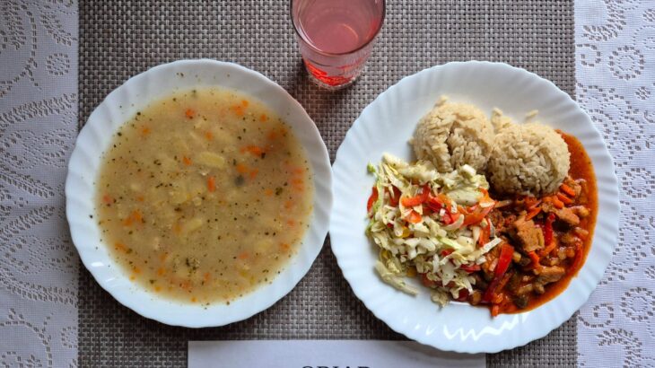 Na zdjęciu znajduję się posiłek w którego skład wchodzi Zupa żurek z ziemniakami, Ryż basmati, Wieprzowina w sosie słodko-kwaśnym, Surówka z kapusty pekińskiej, papryki i kukurydzy oraz kompot z jabłek